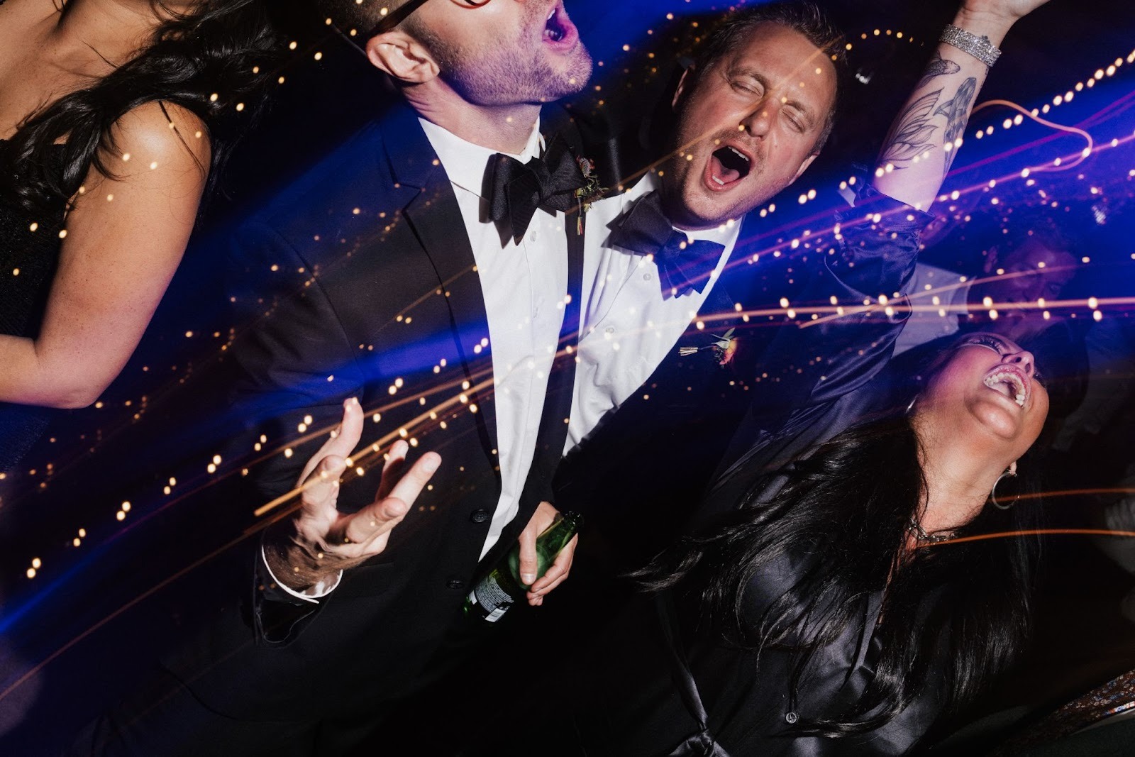 Close dance-floor candid with two guests mid-laugh/cheer under string lights, shot with motion and tight framing. Shot with a 28mm lens by Esteban. 