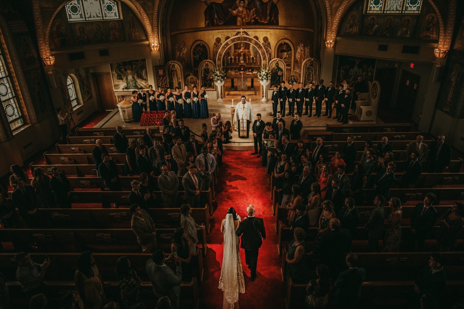 Wide wedding ceremony scene inside a church, photographed from the back with guests seated and the couple at the front. Shot with a 28mm lens by Esteban.