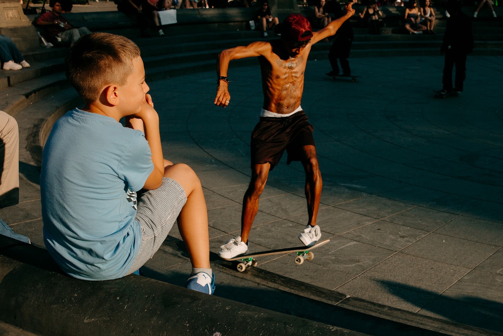 Child seated in the foreground watching a shirtless skateboarder mid-move in an open public space. Shot with a 28mm lens by Esteban. 
