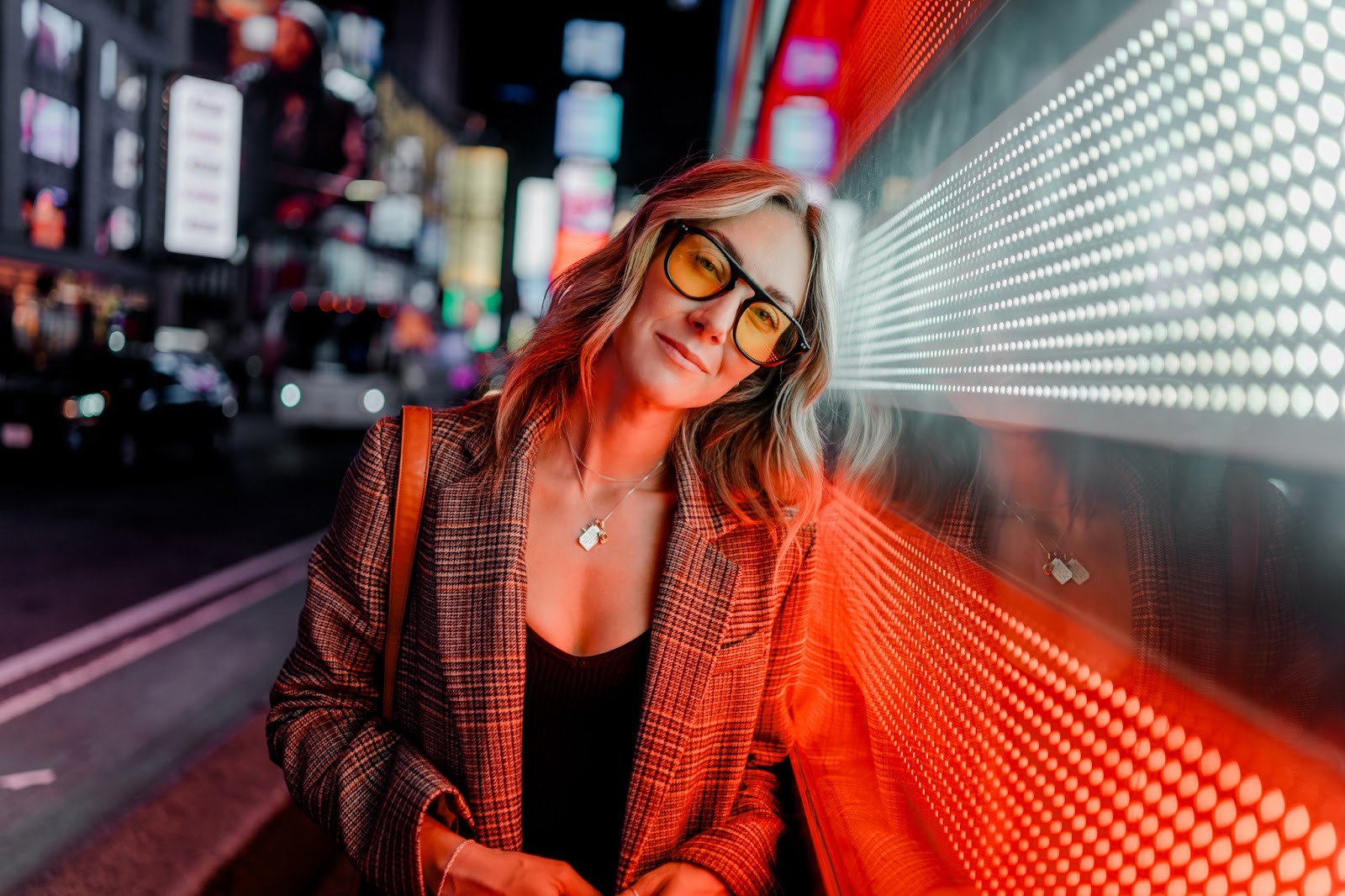 Close portrait of a woman leaning against a bright LED-lit wall at night, with colorful city lights behind her. Shot with a 28mm lens by Esteban. 