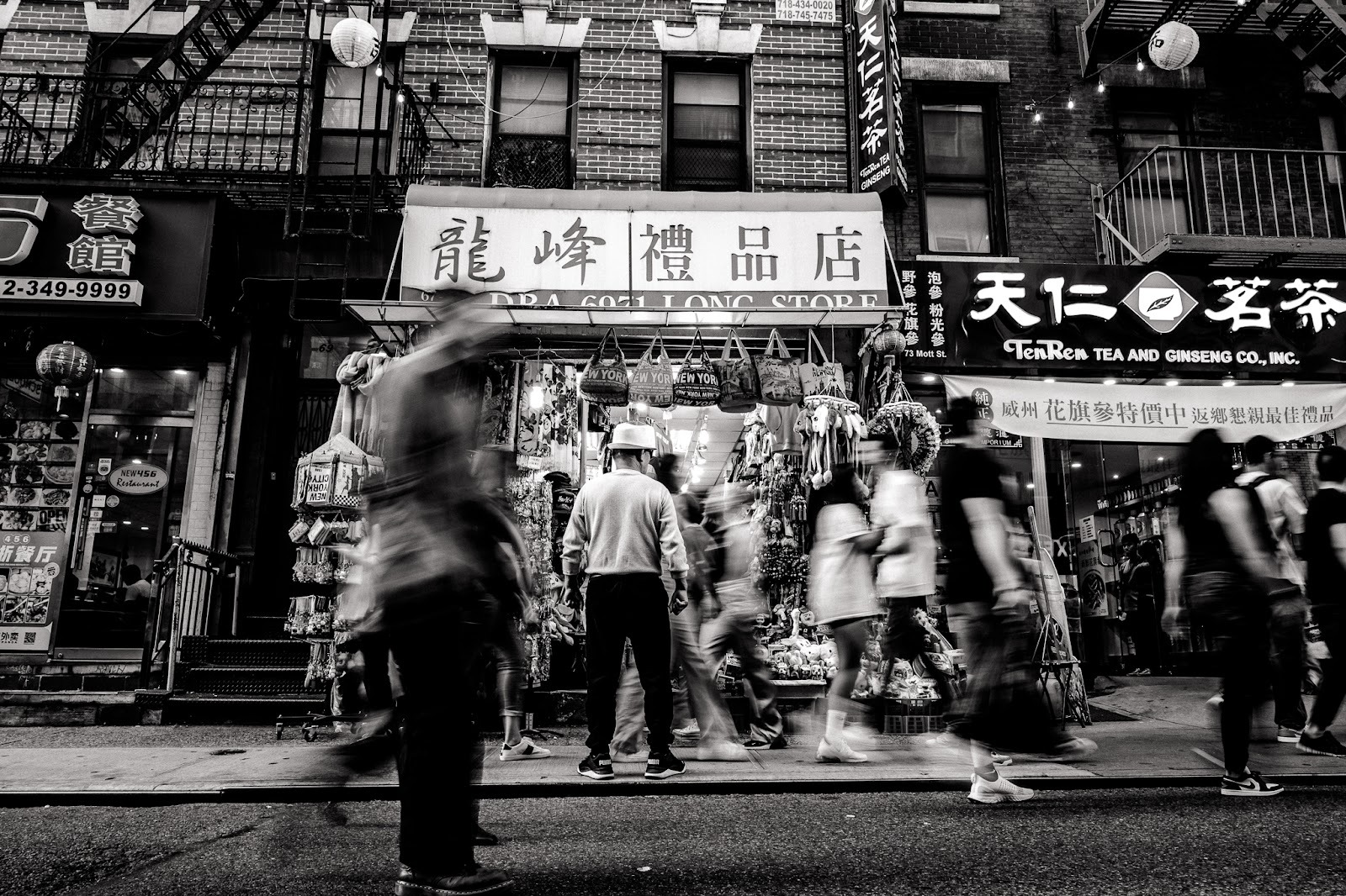 Black-and-white street scene with storefronts and Chinese signage, pedestrians blurred in motion across the frame. Shot with a 28mm lens by Esteban. 