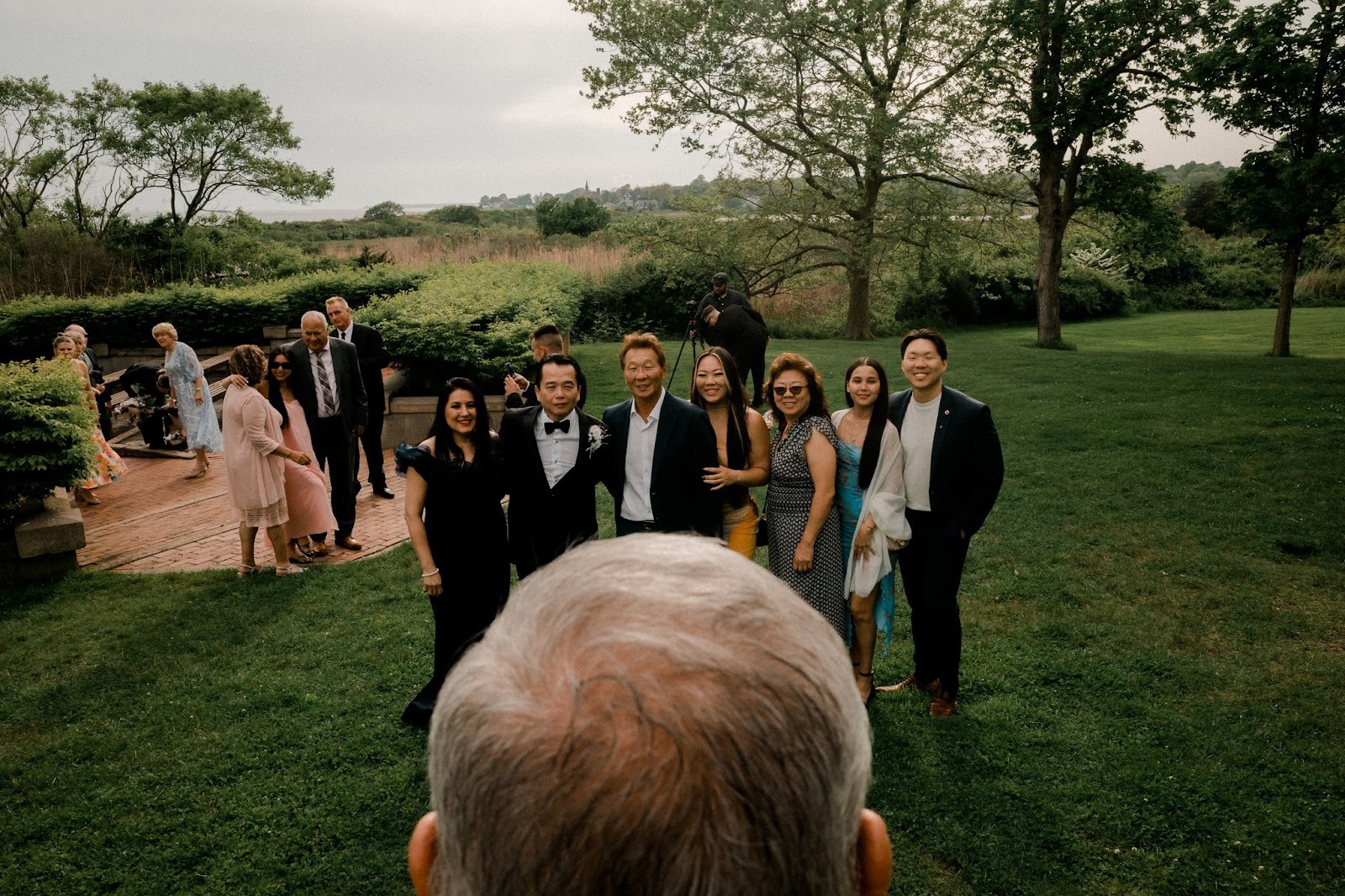 Group of wedding guests posing outdoors on a lawn, photographed from behind a person’s head in the foreground. Shot with a 28mm lens by Esteban.