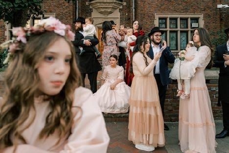 Wedding guests in pale outfits gathered outside a brick building, with a softly blurred person in the foreground. Shot with 28mm lens by Esteban. 
