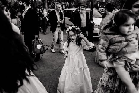 Black-and-white candid of a child in a dress walking through a crowd, with motion and people partially cut at the frame edges. Shot with a 28mm lens by Esteban.